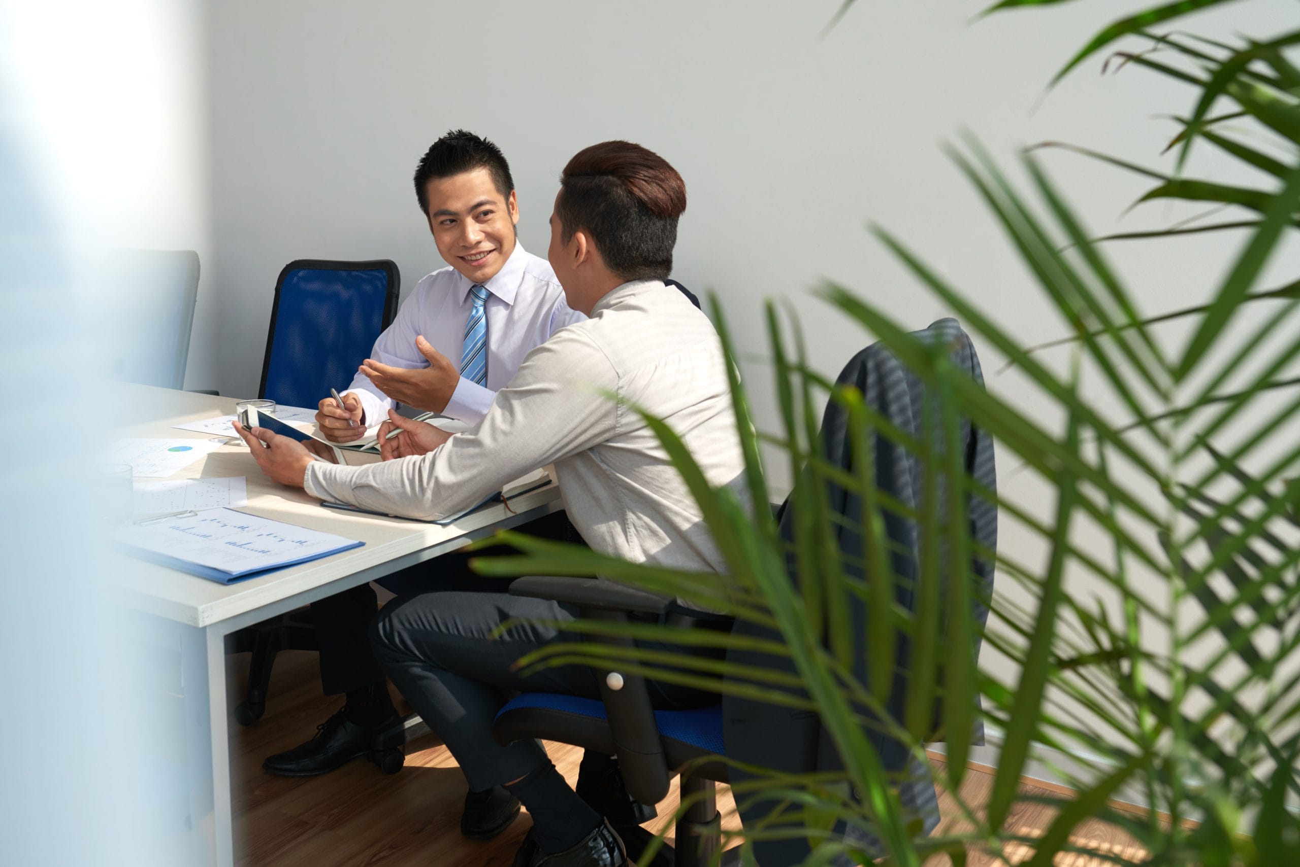 Two men in office discussing at a desk with papers and a pen, plant leaves framing the scene.