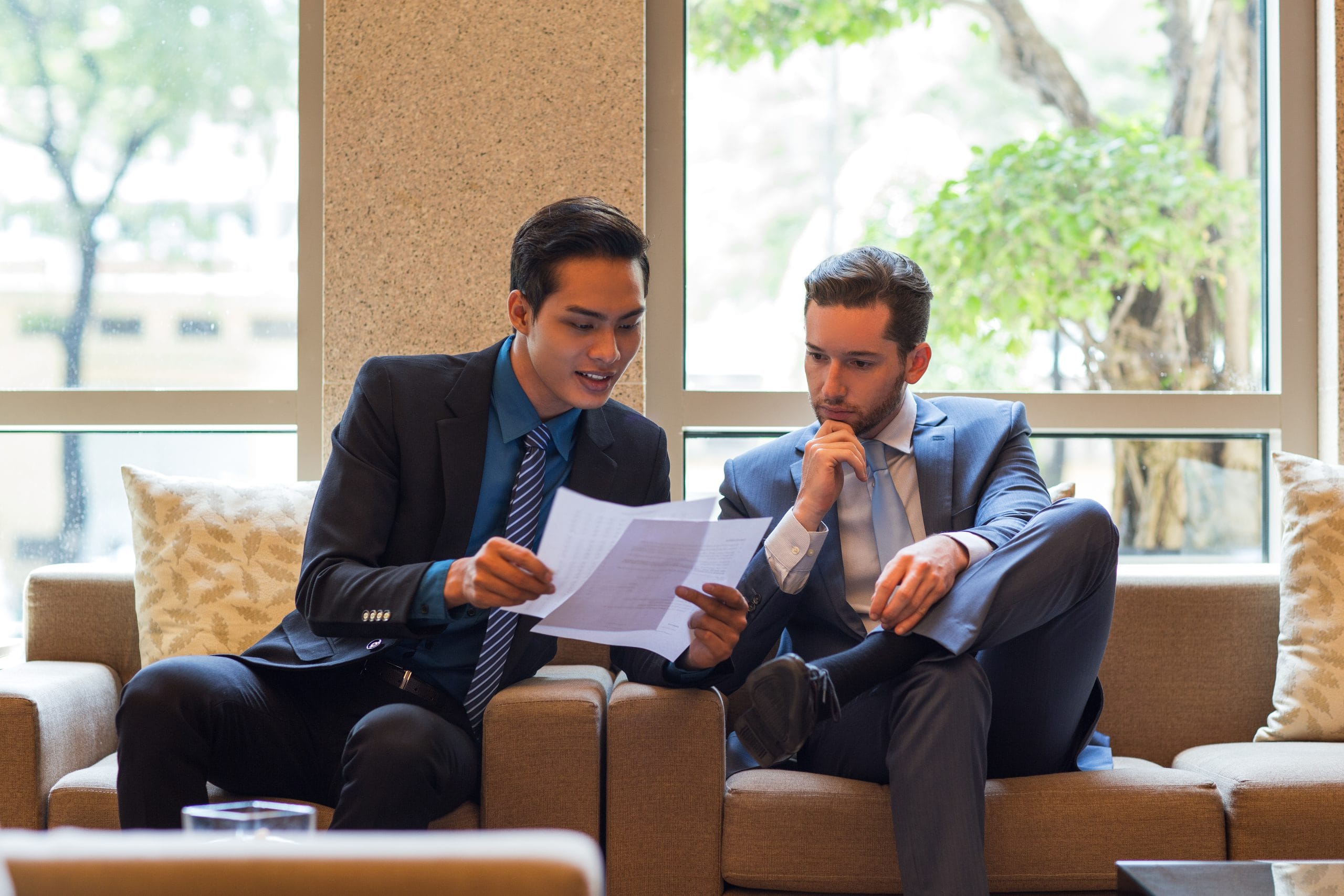 Two men in business suits sit on a beige sofa, reviewing printed documents together in a bright lounge with large windows behind them.
